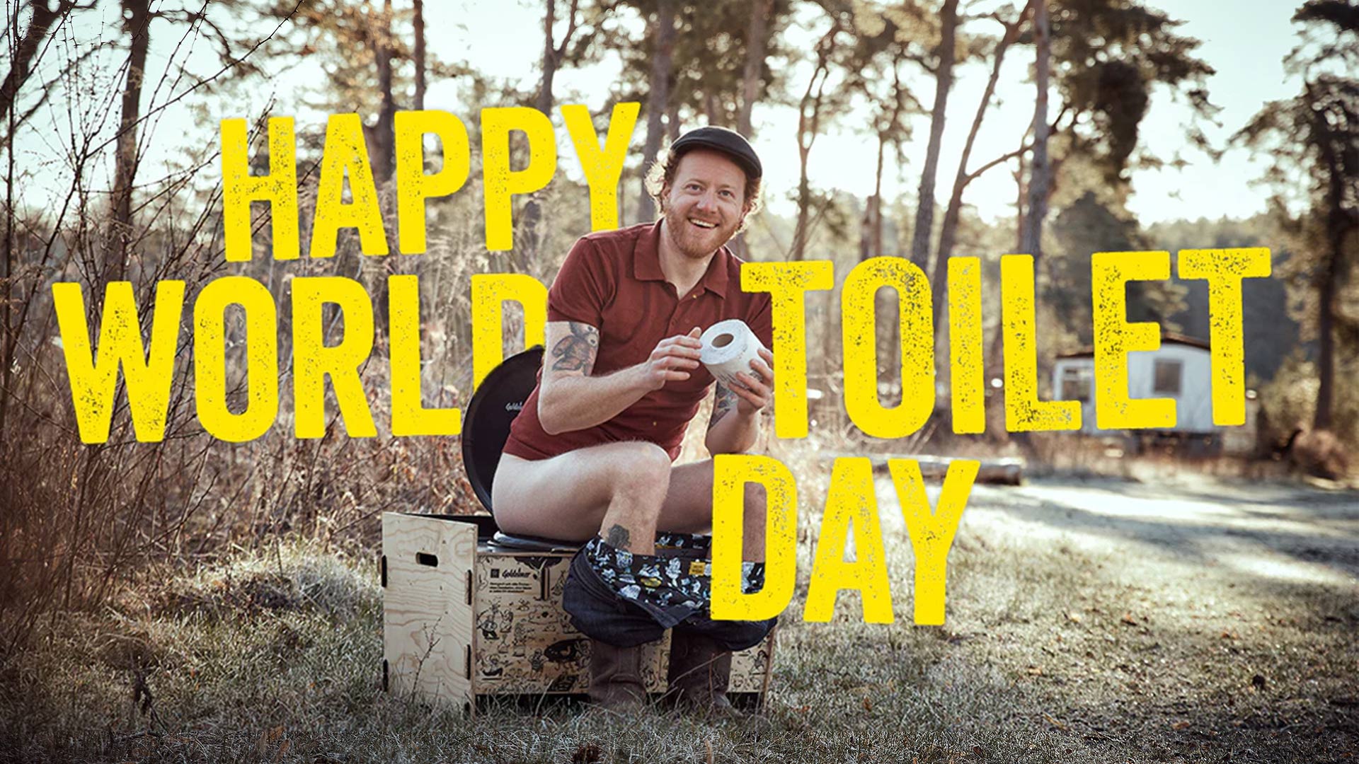 Mann sitzt lachend auf Trockentoilette, großer Schriftzug "Happy World Toilet Day"