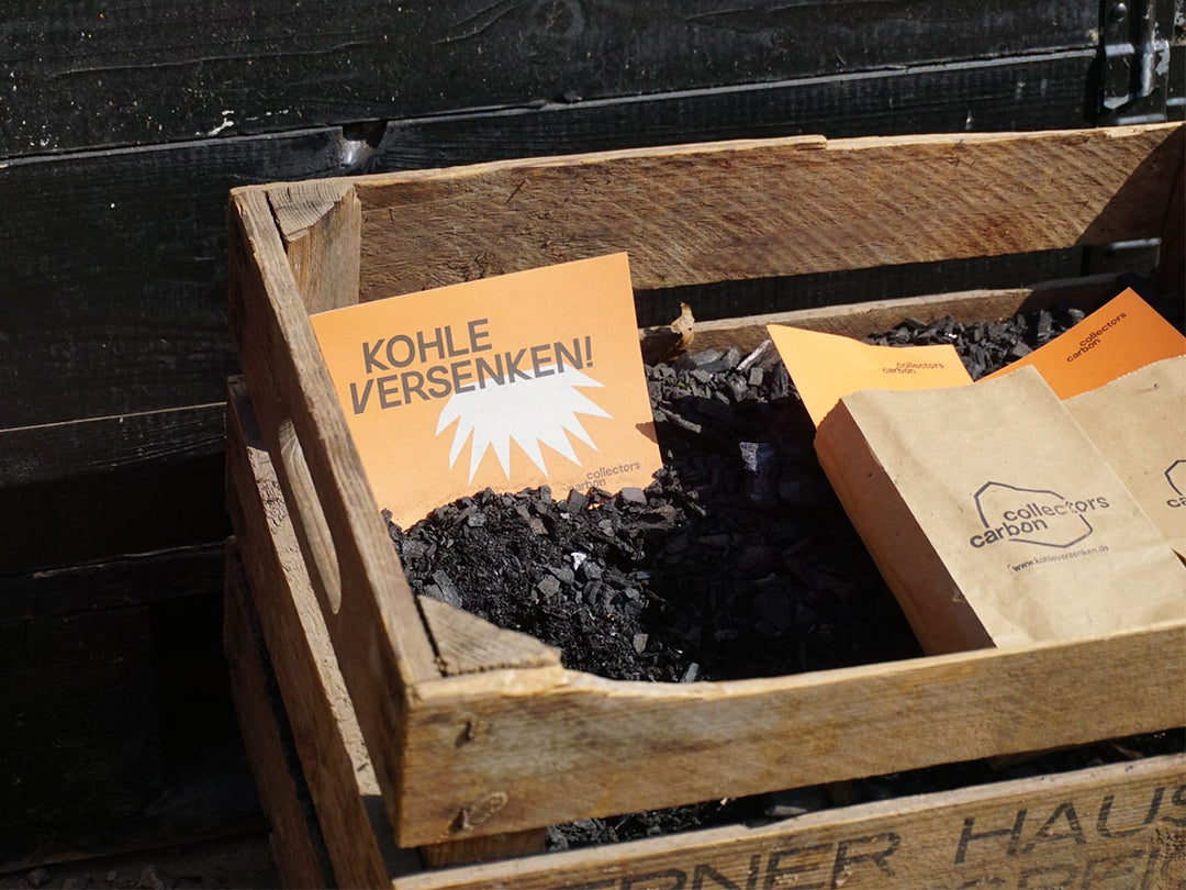 Holzbox mit Pflanzenkohle, darin liegt ein Flyer, auf dem "Kohle versenken!" steht