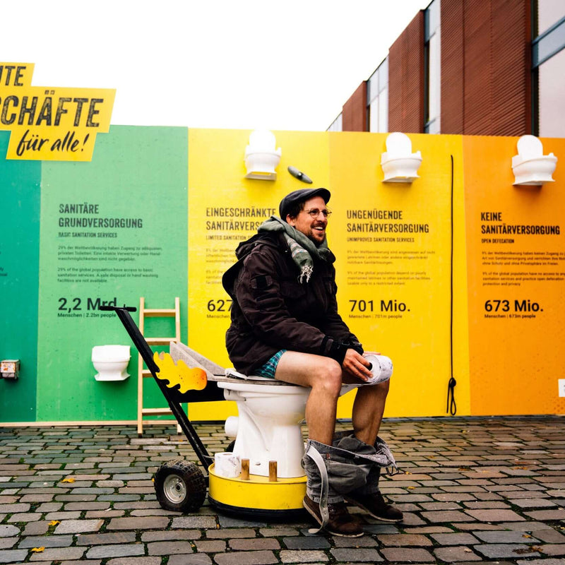 Malte sitzt mit heruntergelassener Hose auf einer Toilette vor der bunten Sanitation Ladder Wand, auf der die Stufen der Sanitärversorgung erklärt sind.