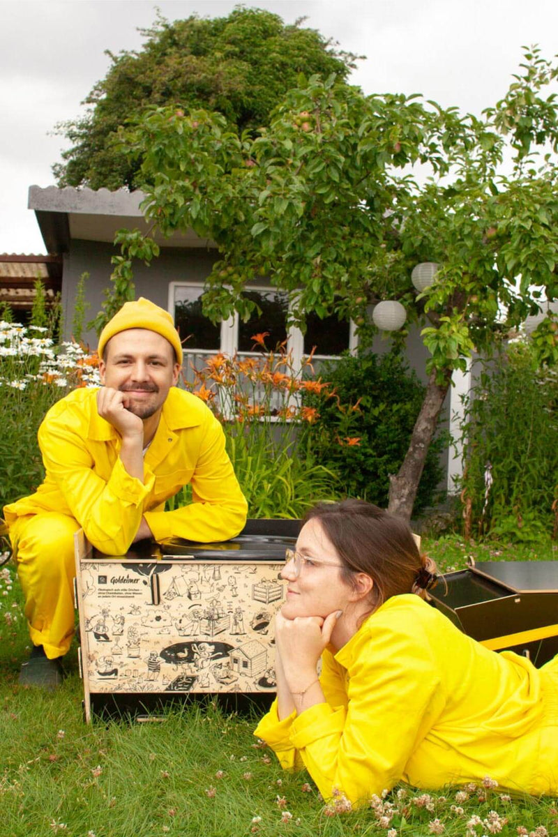 Manni und Tanja in gelben Overalls im Garten vor der Goldeimer Premium Toilette